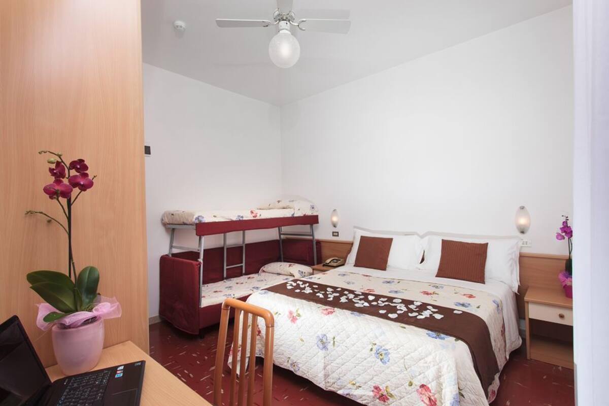 A bedroom with a bed, a desk with a laptop, a chair, a ceiling fan, and a potted plant.