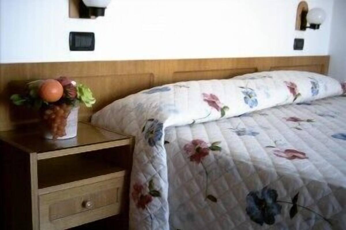 A bedroom with a wooden nightstand, a drawer, a vase with flowers, and a floral-patterned bedspread.