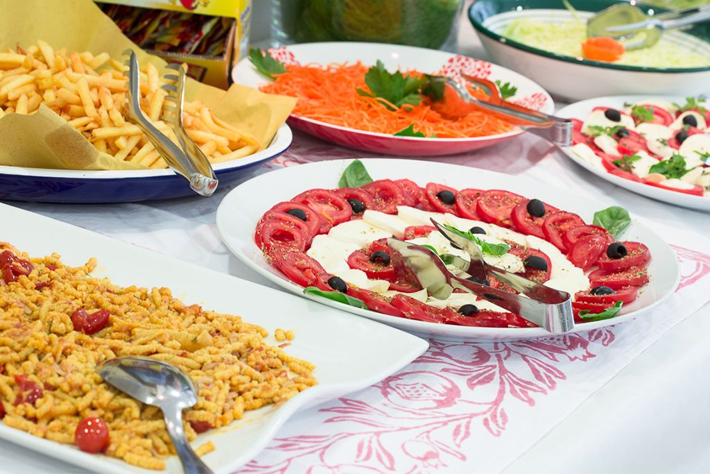A table with various dishes including a salad with tomatoes, olives, and cheese, a plate of pasta, and a bowl of soup.