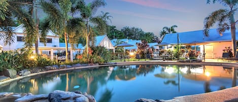 A pool area with a clear water surface, surrounded by rocks and palm trees, with buildings in the background.