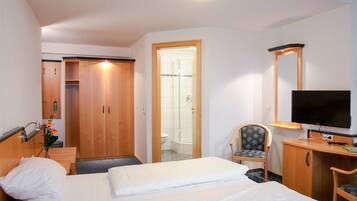 A hotel room with a bed, a chair, a desk with a TV, a wardrobe, and a shower area.