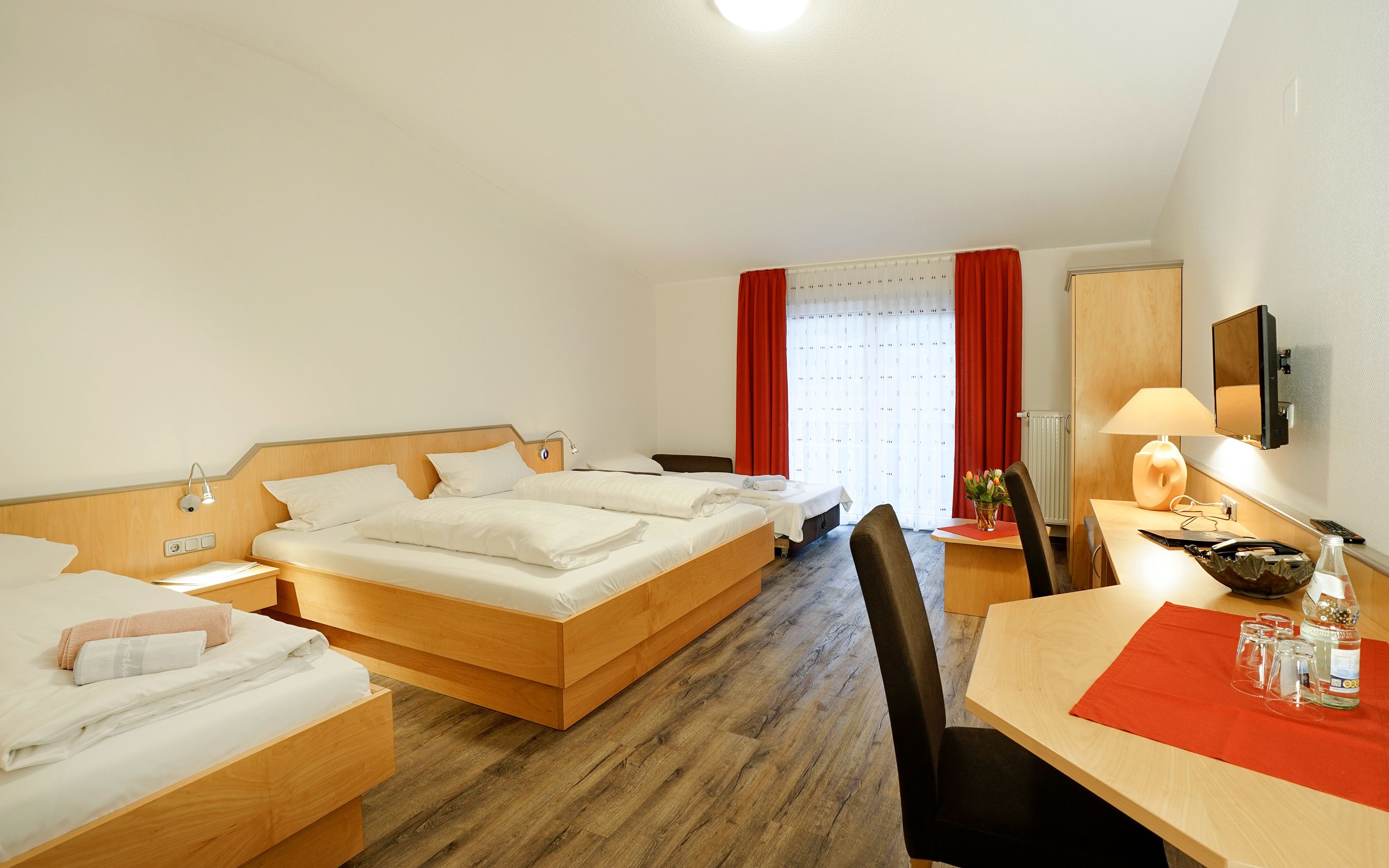 A hotel room with two single beds, a desk with a chair, a TV, and a window with red curtains.