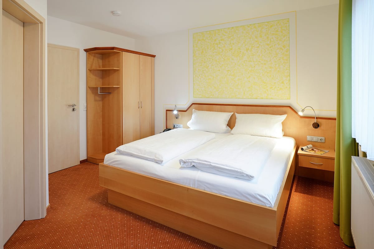 Ein Hotelzimmer mit einem Doppelbett, einem Holzschrank, einer Duschkabine und einem Nachttisch mit Lampe.