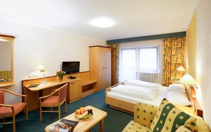 A hotel room with a bed, a desk with a computer, a chair, a small table with fruit, and a television.