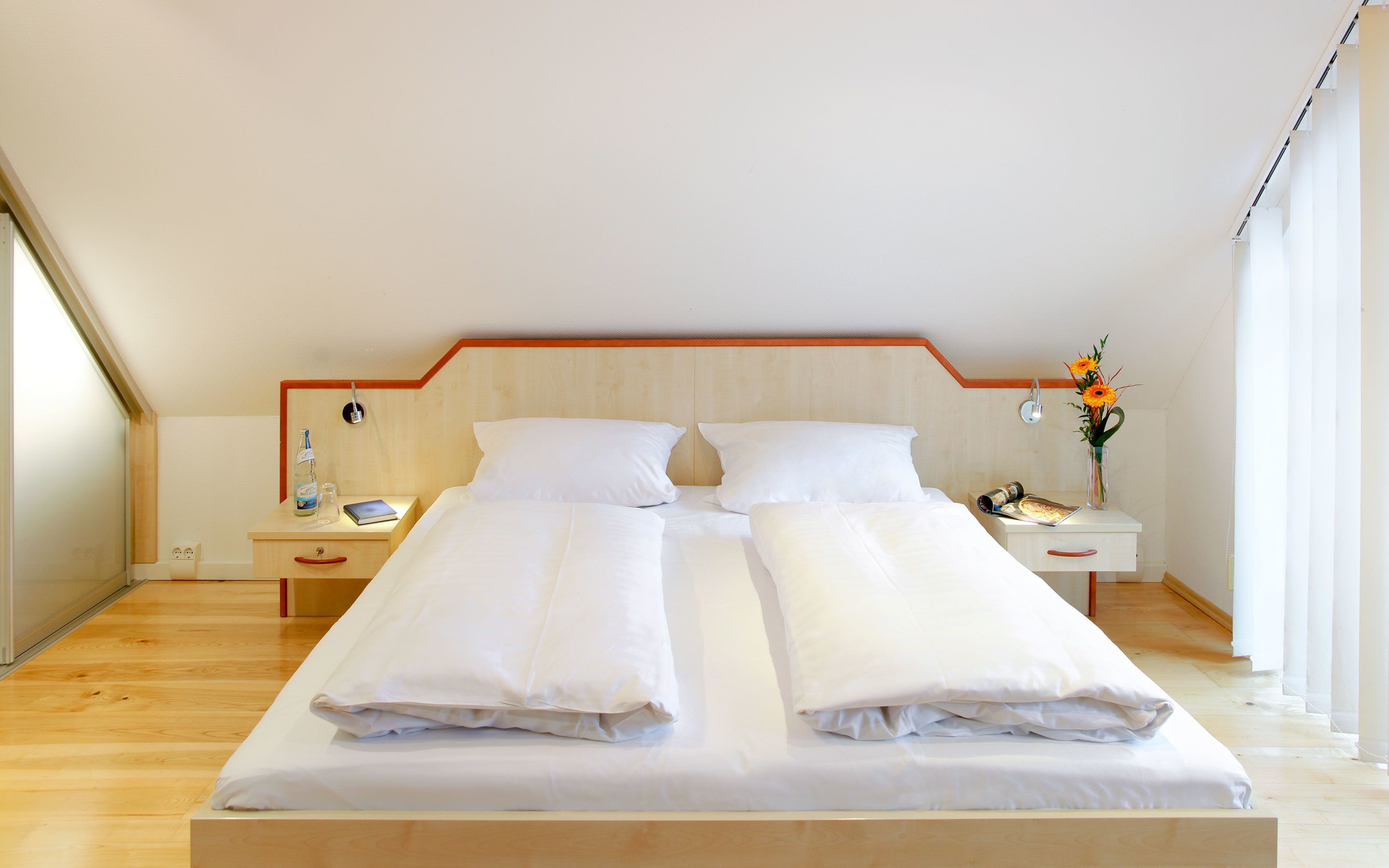 A double bed with white linens, a wooden headboard, a bedside table with a lamp, and a vase with flowers.