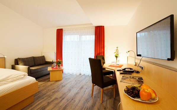 A hotel room with a bed, a sofa, a desk with a computer, a TV, and a dining table with fruit.