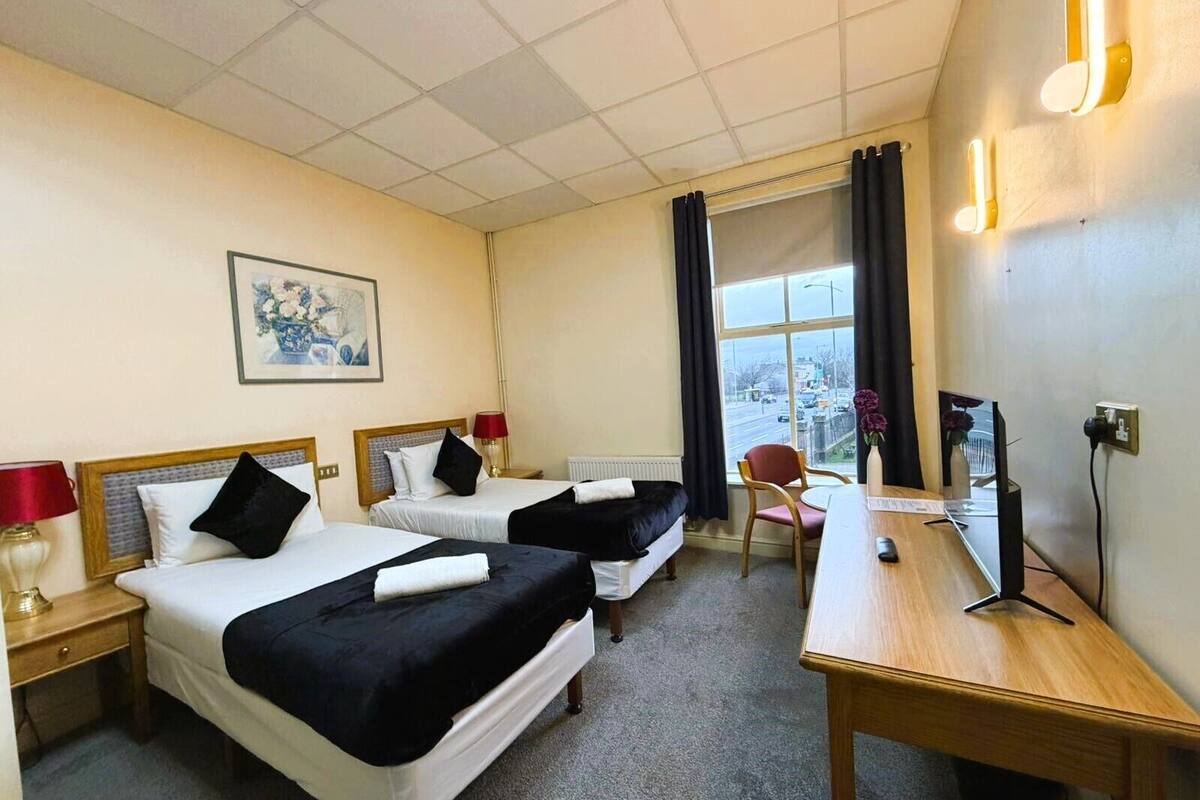 A hotel room with two beds, a desk, a chair, a window with curtains, and a painting on the wall.