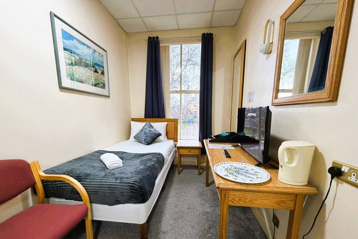 A small, well-lit room with a bed, a desk, a chair, a television, and a window with curtains.