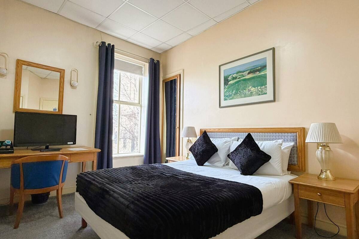 A hotel room with a bed, a desk with a computer, a chair, a lamp, and a painting on the wall.