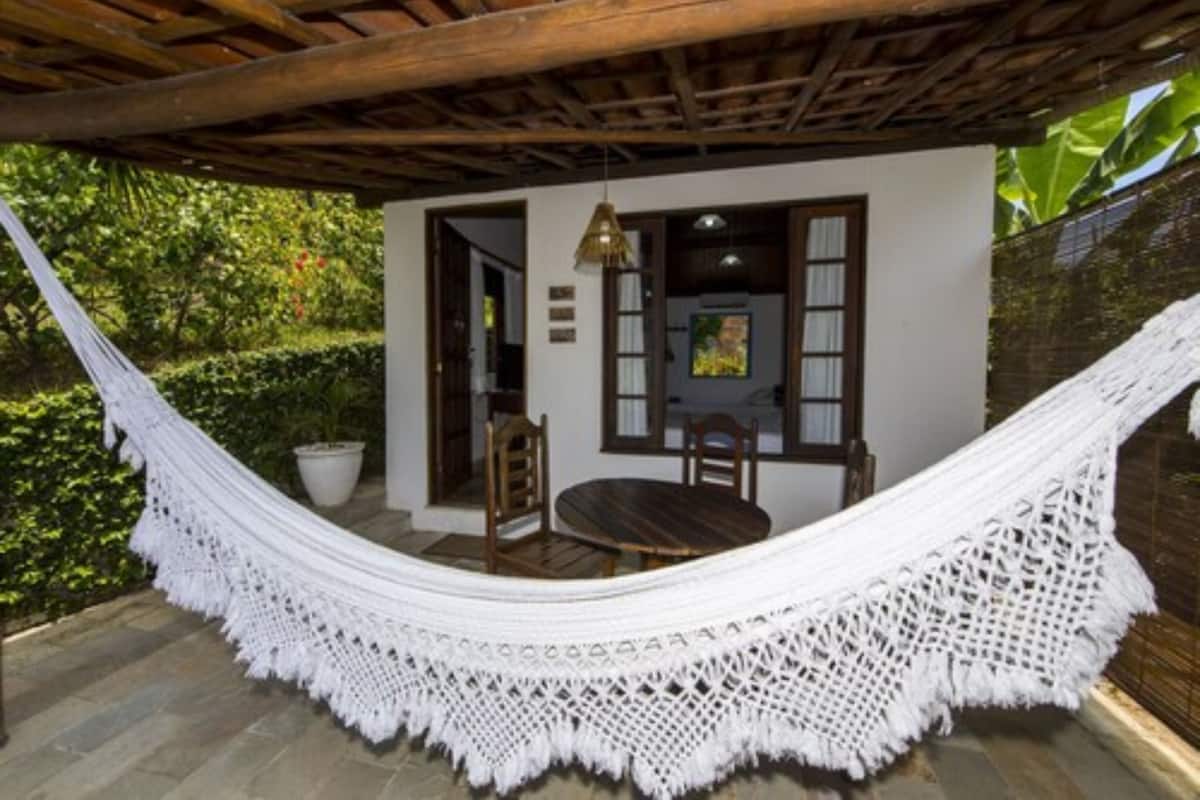 Un hamac sous une pergola en bois, une table en bois avec des chaises, et un mur blanc avec une fenêtre et un tableau.