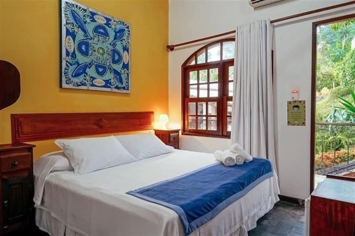 Une chambre d’hôtel avec un lit, une table de chevet, une fenêtre avec des rideaux et une vue sur la verdure.