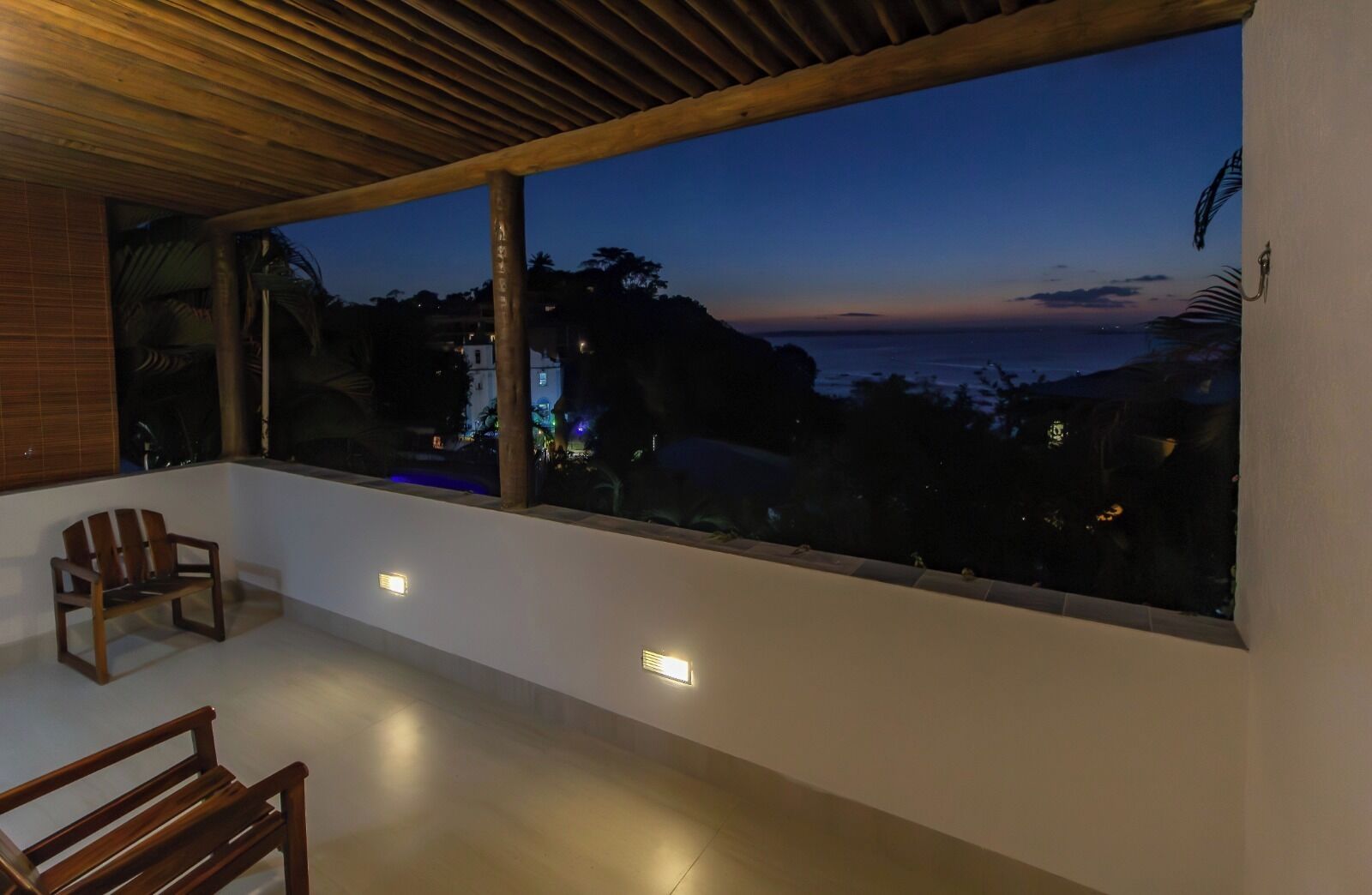 Varanda com vista para o oceano ao entardecer, mobiliário de madeira e iluminação embutida.
