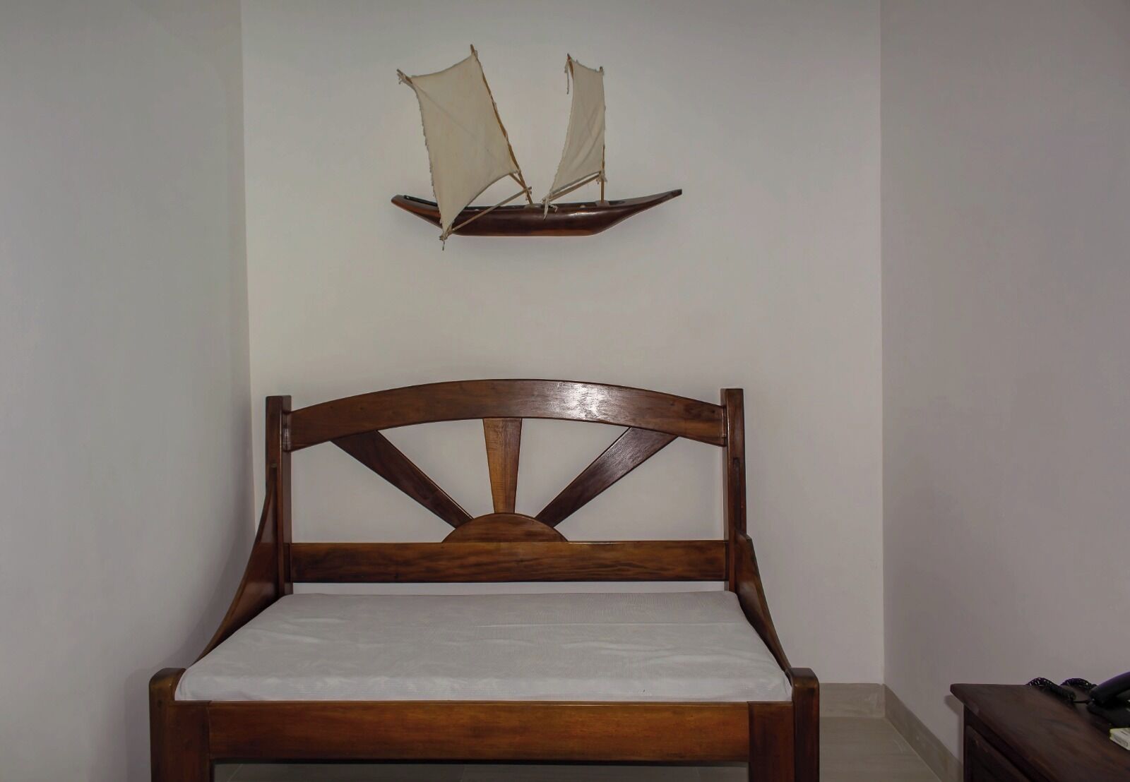 Un lit en bois avec une tête de lit, une étagère fixée au mur ornée d’une maquette de voilier, et une petite table de chevet avec une lampe.