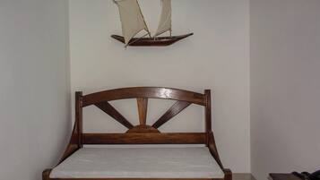 Un lit en bois avec une tête de lit, une étagère fixée au mur ornée d’une maquette de voilier, et une petite table de chevet avec une lampe.