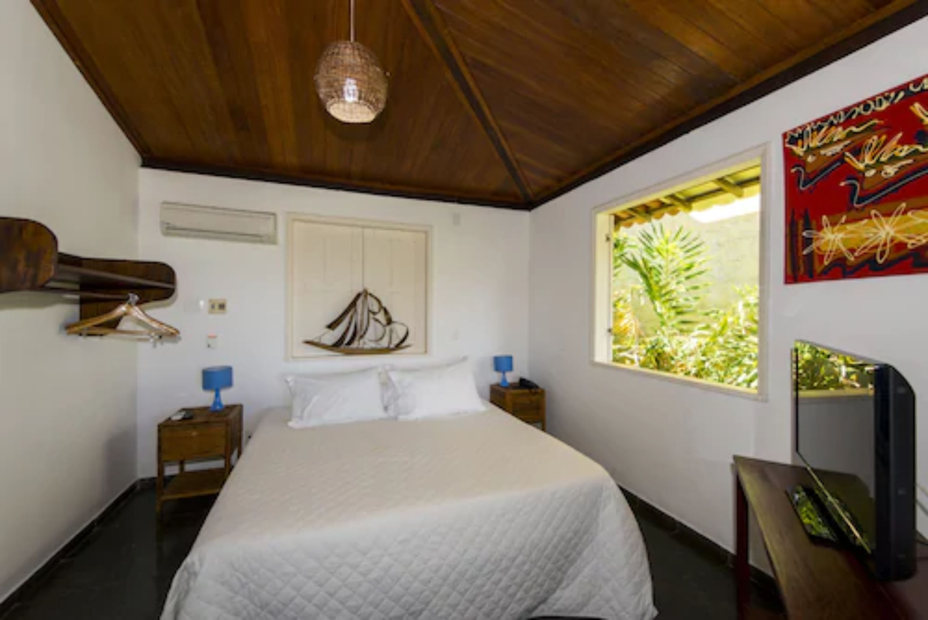 Un dormitorio con techo de madera, una cama con colcha blanca, una mesita de noche con lámpara, un televisor montado en la pared y una ventana con vistas a zonas verdes.