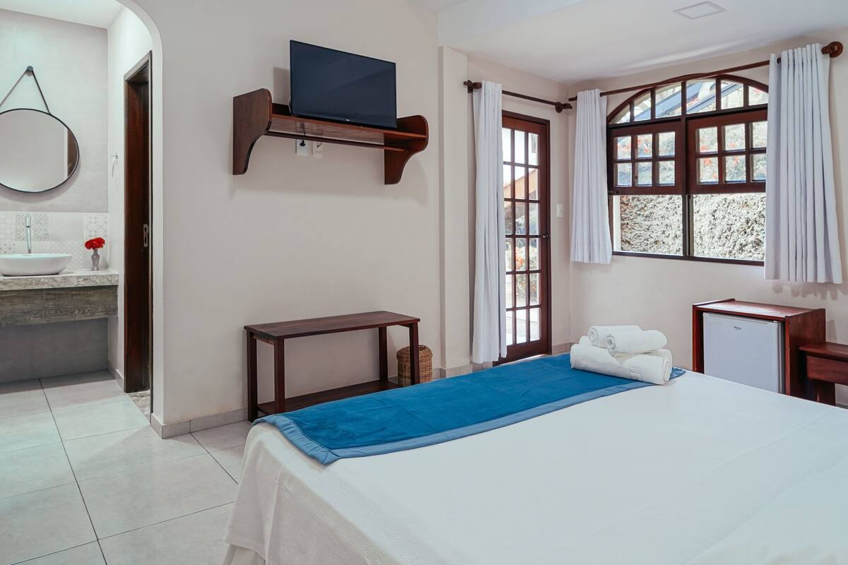 Une chambre d’hôtel avec un lit, un téléviseur posé sur une étagère, une fenêtre avec des rideaux, une petite table et un bureau avec une lampe.