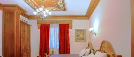 A bedroom with two beds, wooden headboards, a chandelier, a painting, and a red curtain.