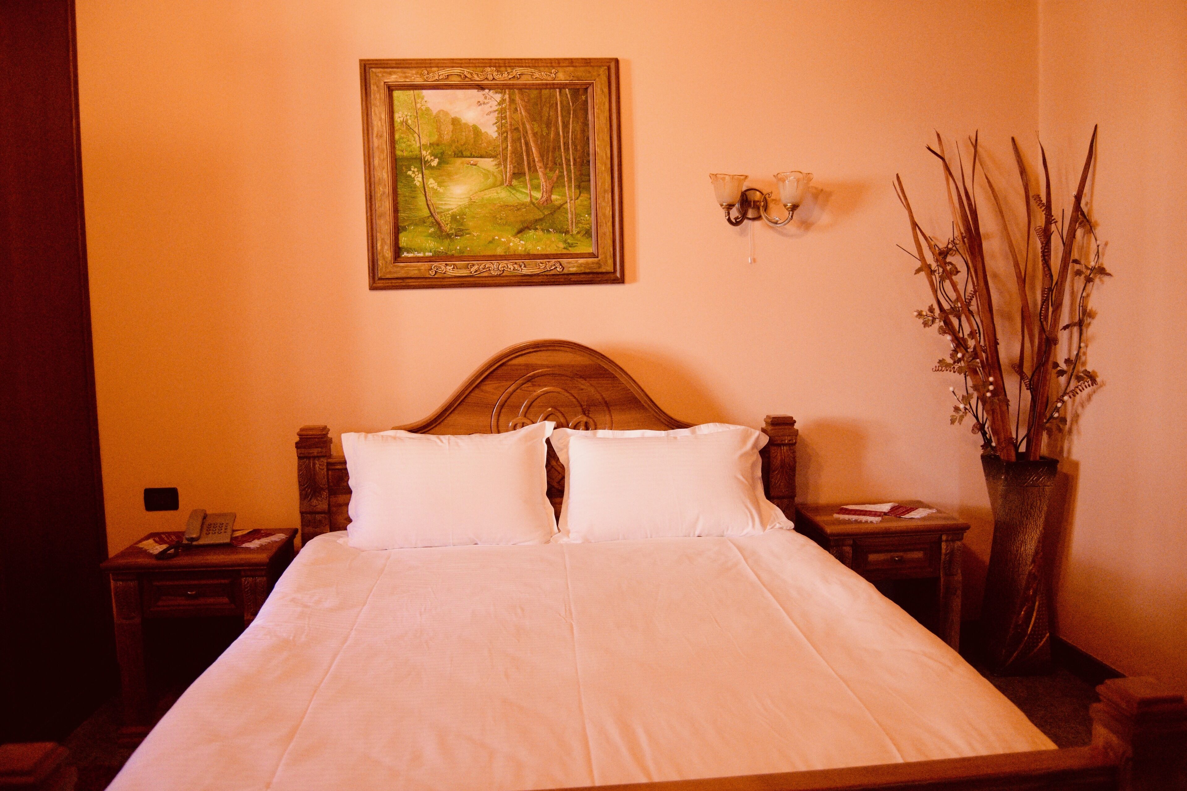 A neatly made bed with white linens, a wooden headboard, a bedside table with a lamp, a wall-mounted painting of a forest, and a vase with branches.