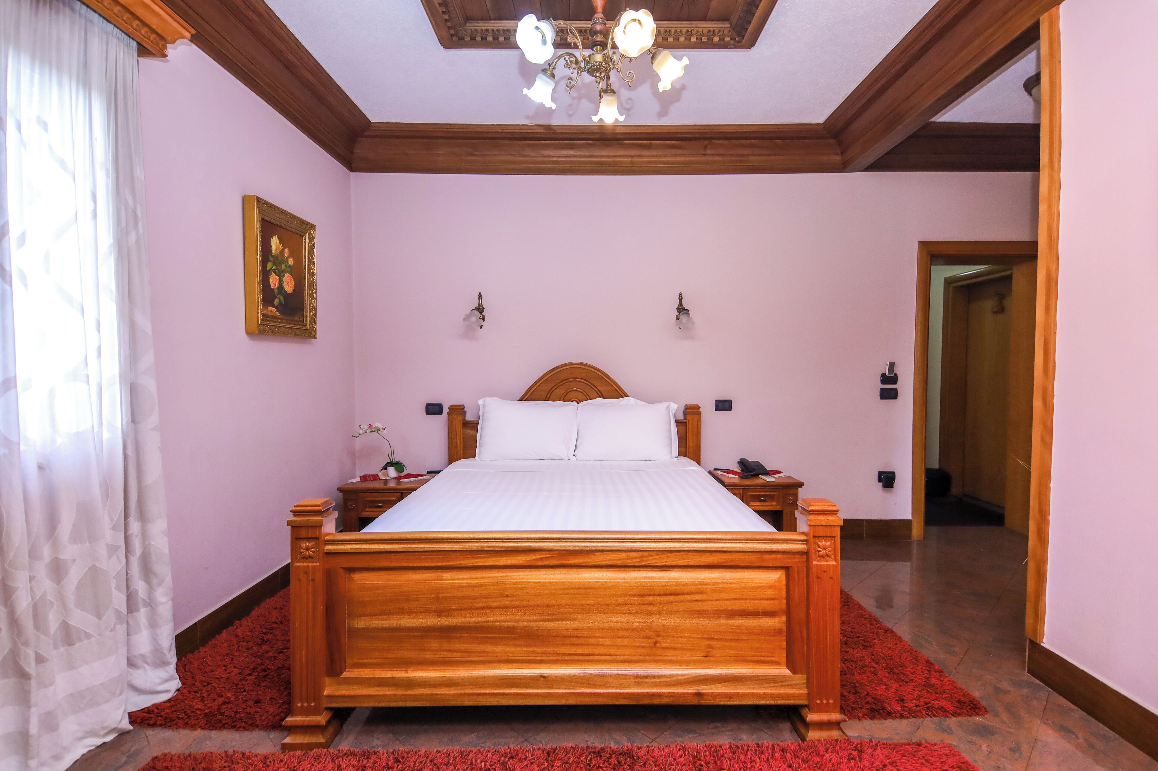 A bedroom with a wooden bed, a chandelier, a painting on the wall, and a red rug.