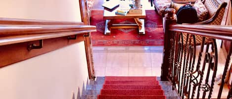 Una escalera conduce a una sala de estar con alfombra roja, barandillas de madera y un candelabro.