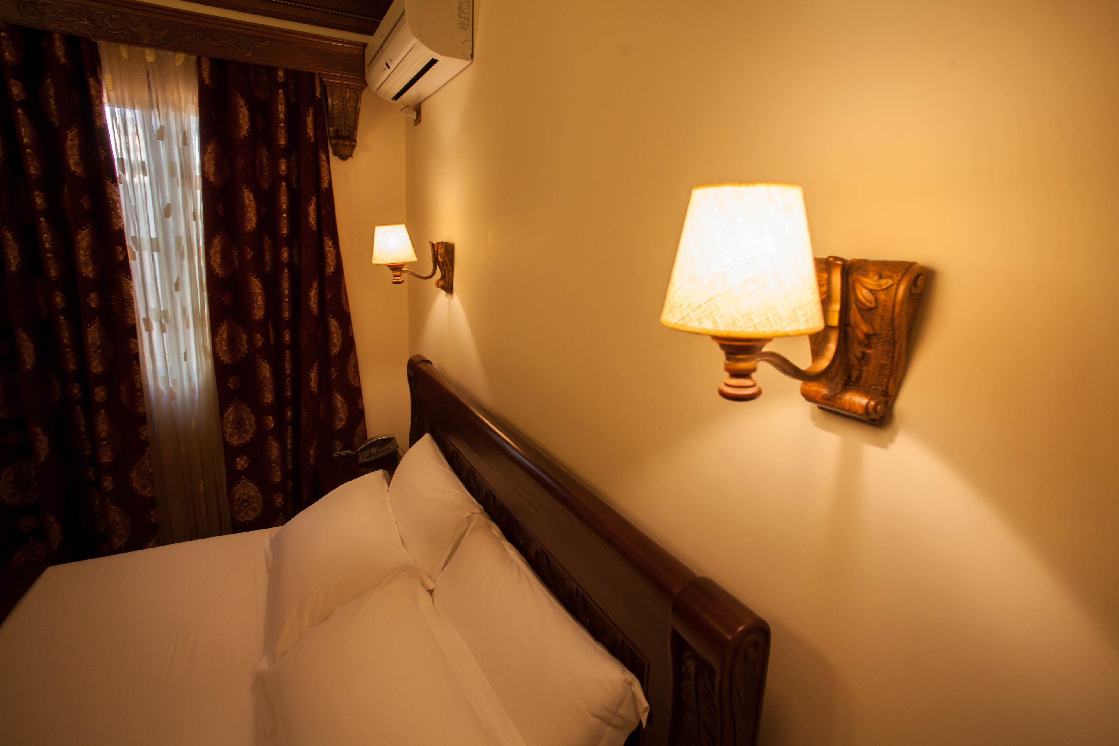 A hotel room with a bed, a wall-mounted lamp, and an air conditioning unit.