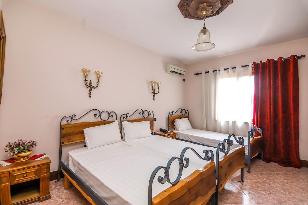 A bedroom with two beds, wooden headboards, a nightstand with a lamp, and a window with red curtains.