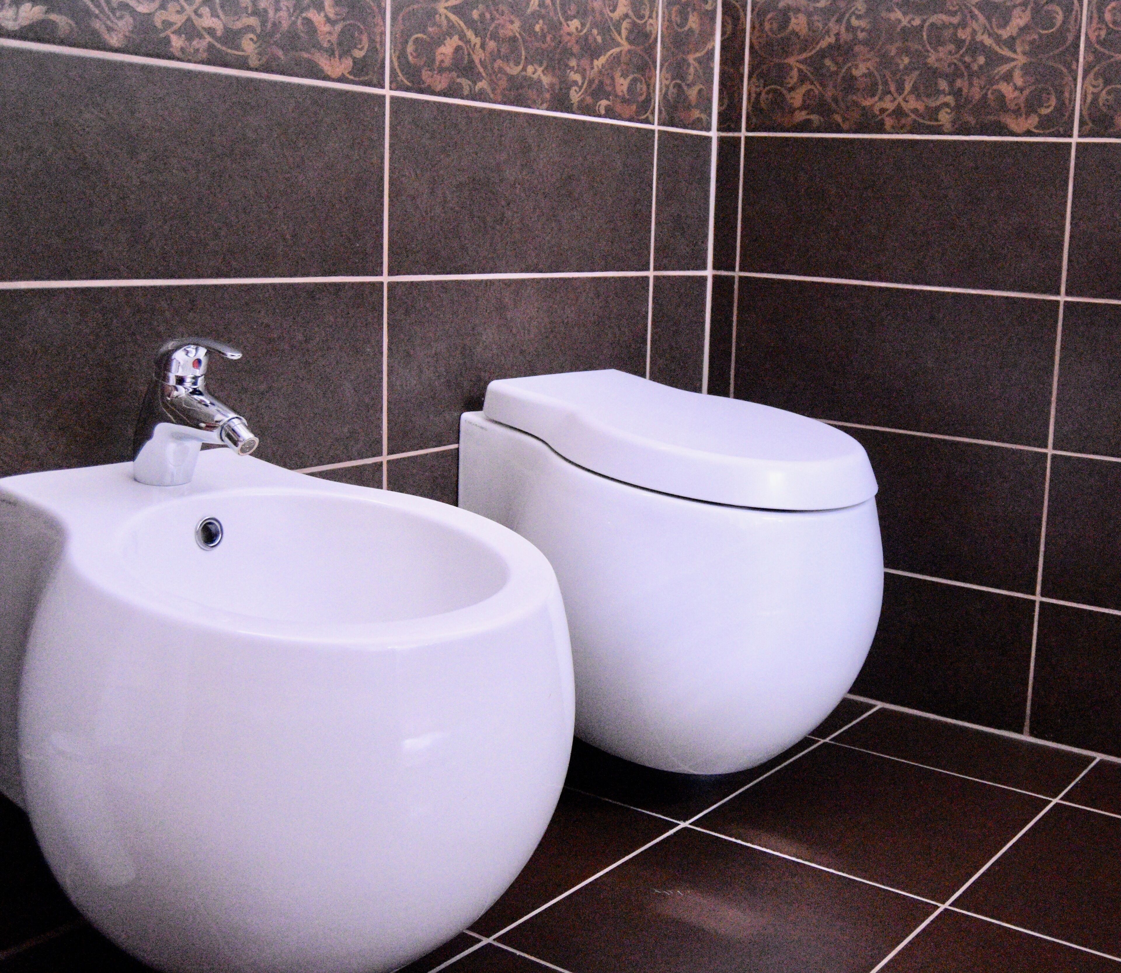 A bathroom with a bidet, a toilet, and dark patterned tiles.