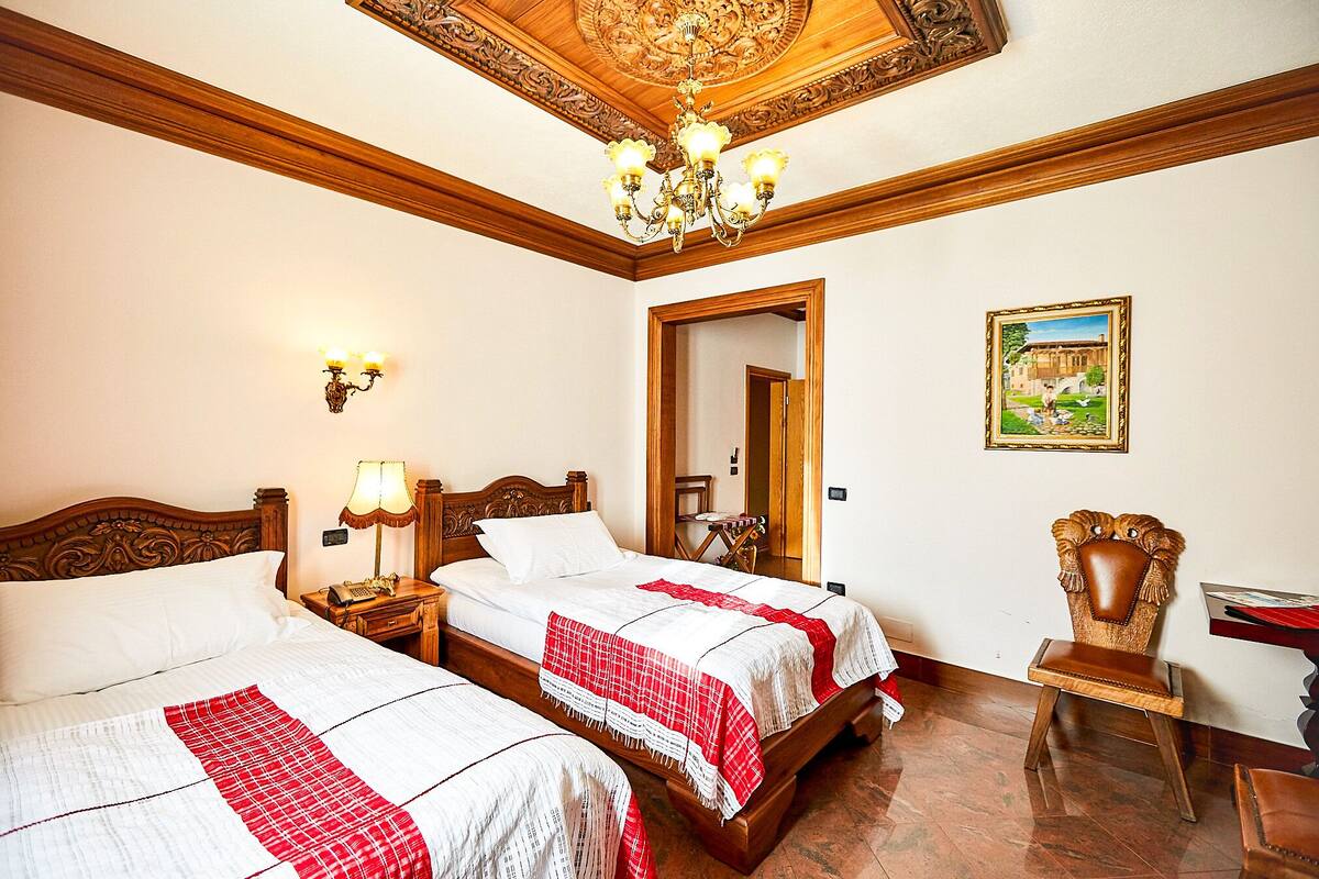 A hotel room with two beds, wooden headboards, a chandelier, a painting, and a wooden chair.