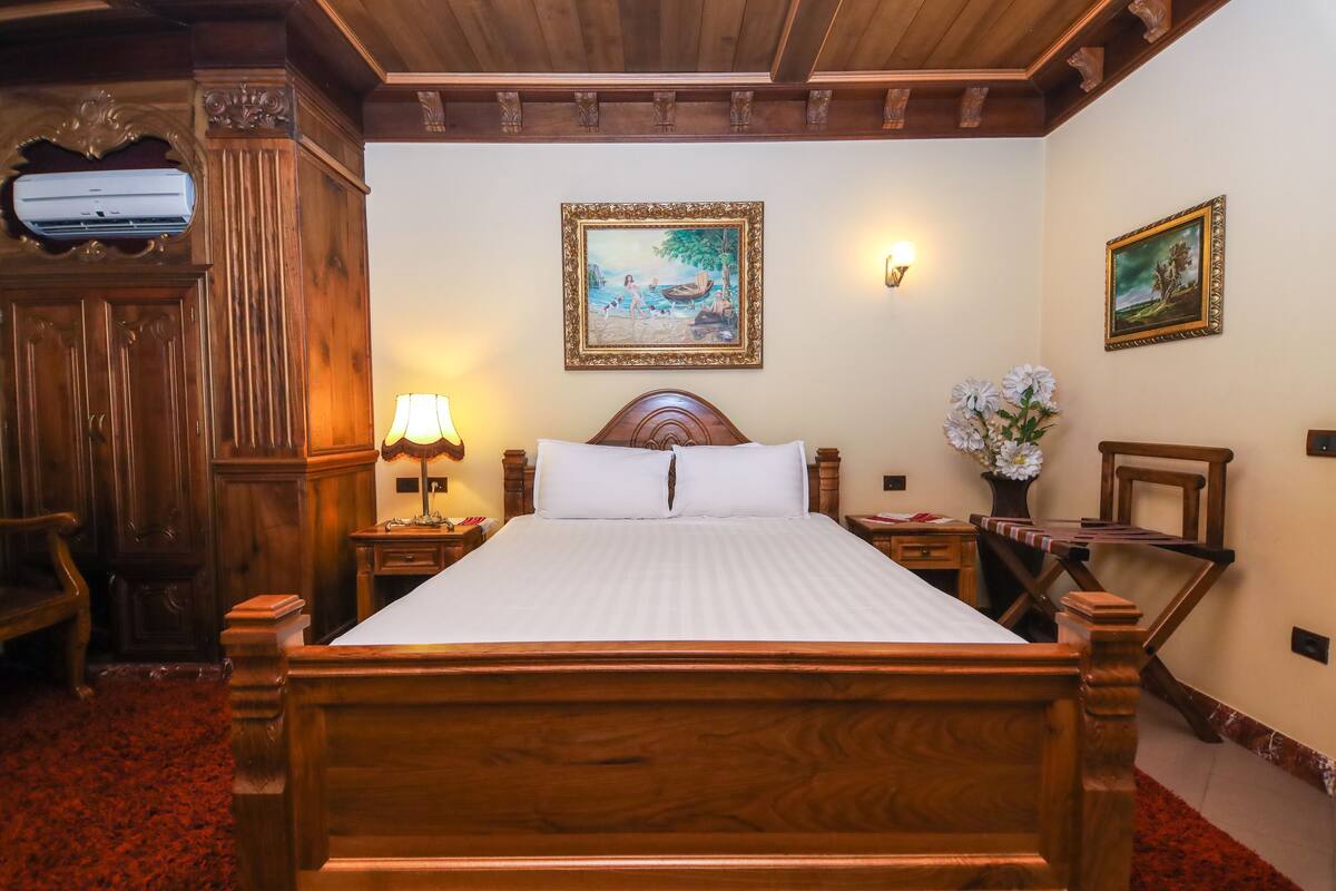 A wooden four-poster bed with white linens, a nightstand with a lamp, a wooden chair, and two framed paintings on the wall.