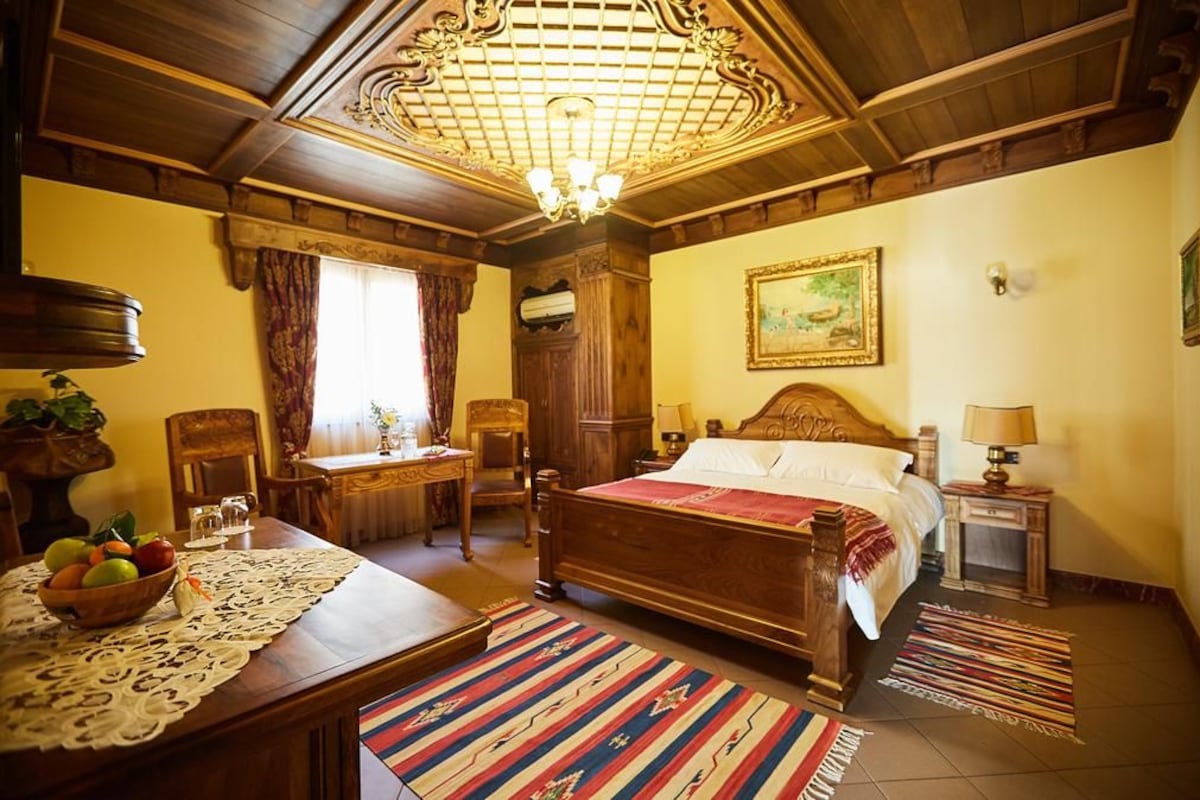 A traditional room with a wooden bed, a dining table with a lace tablecloth, a chandelier, and a painting on the wall.