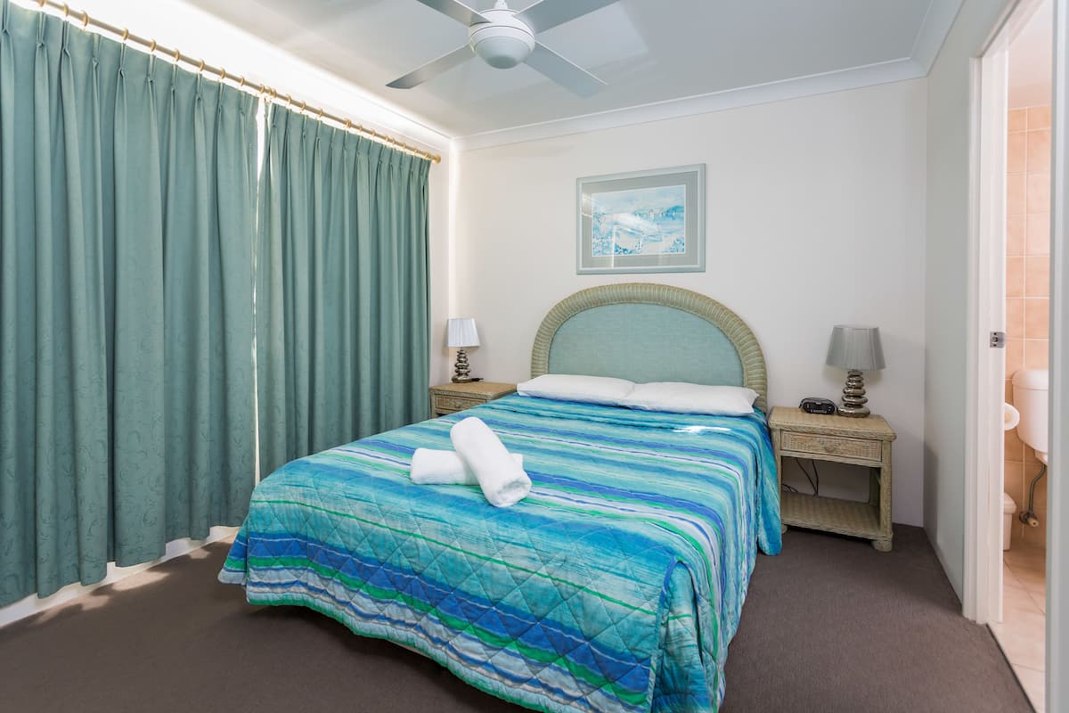 A hotel room with a bed, two bedside tables, a ceiling fan, a framed picture on the wall, and a bathroom door.