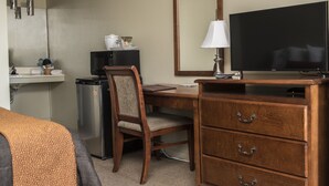A hotel room with a wooden desk, a television, a small refrigerator, a microwave, a mirror, and a bedside table with a lamp. - Motel Le Deauville (Trois-Rivieres)