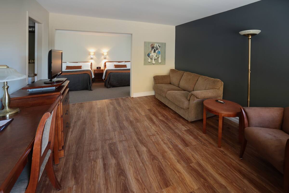 A hotel room with a wooden floor, a sofa, a chair, a desk with a lamp, a TV, and a painting on the wall.
