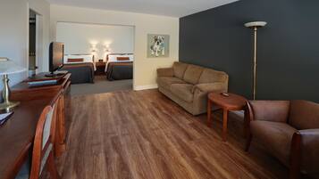 Et hotelværelse med trægulv, en sofa, en lænestol, et skrivebord med lampe, et fjernsyn og et maleri på væggen.