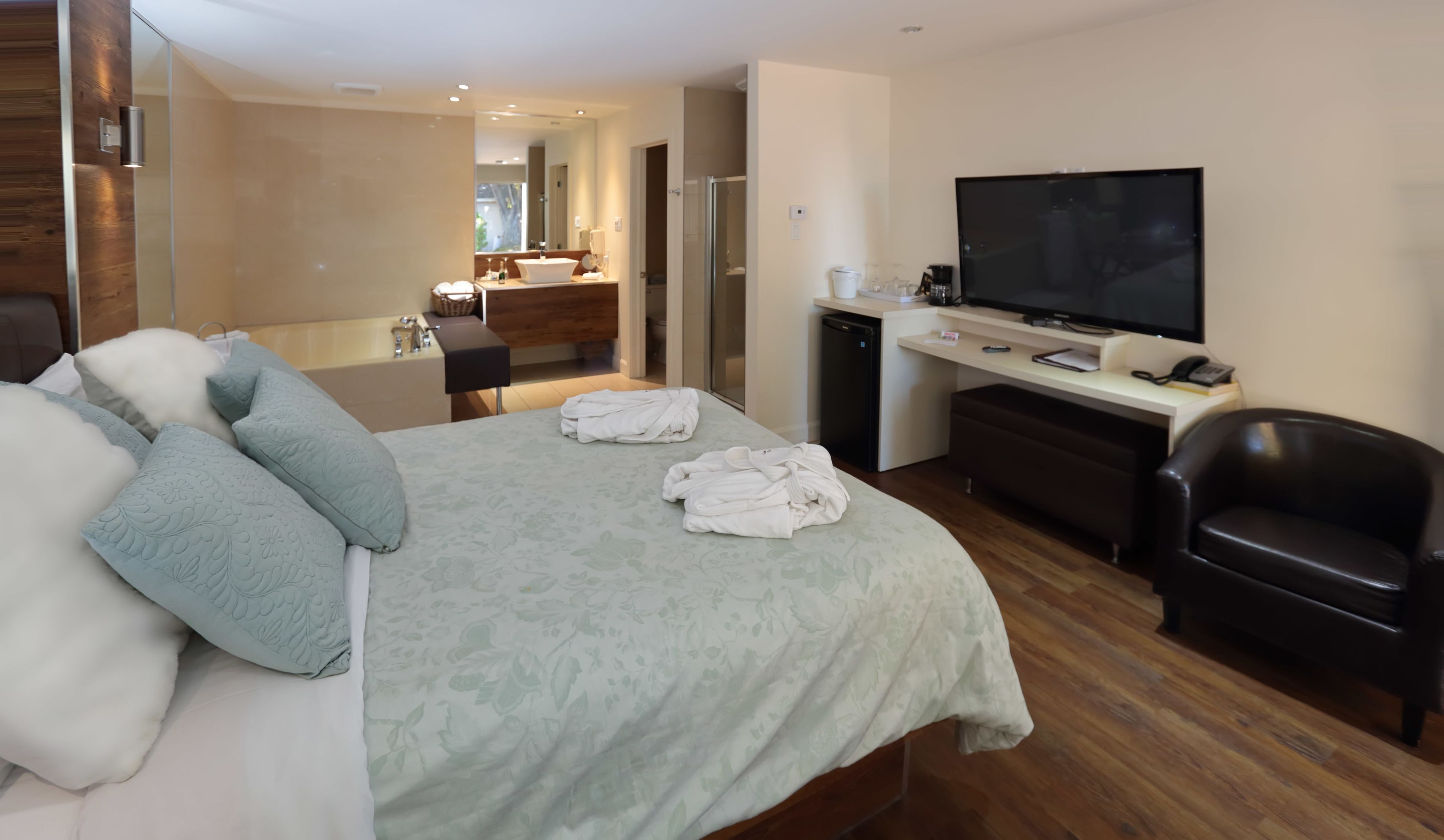 Ein modernes Hotelzimmer mit einem großen Bett, einem Schreibtisch mit Flachbildfernseher, einem Ledersessel und einem durch eine geöffnete Tür einsehbaren Badezimmer.