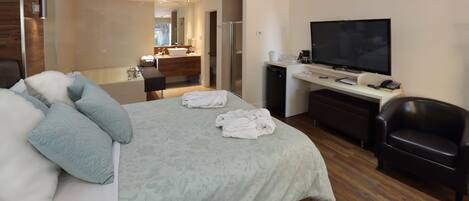Ein modernes Hotelzimmer mit einem großen Bett, einem Schreibtisch mit Flachbildfernseher, einem Ledersessel und einem durch eine geöffnete Tür einsehbaren Badezimmer.