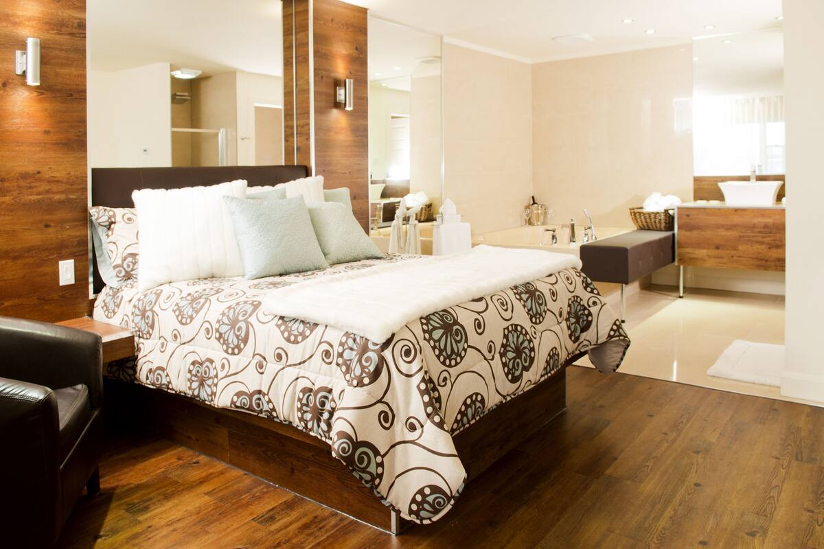 A modern bedroom with a large bed, a wooden headboard, a nightstand, and a bathroom in the background.