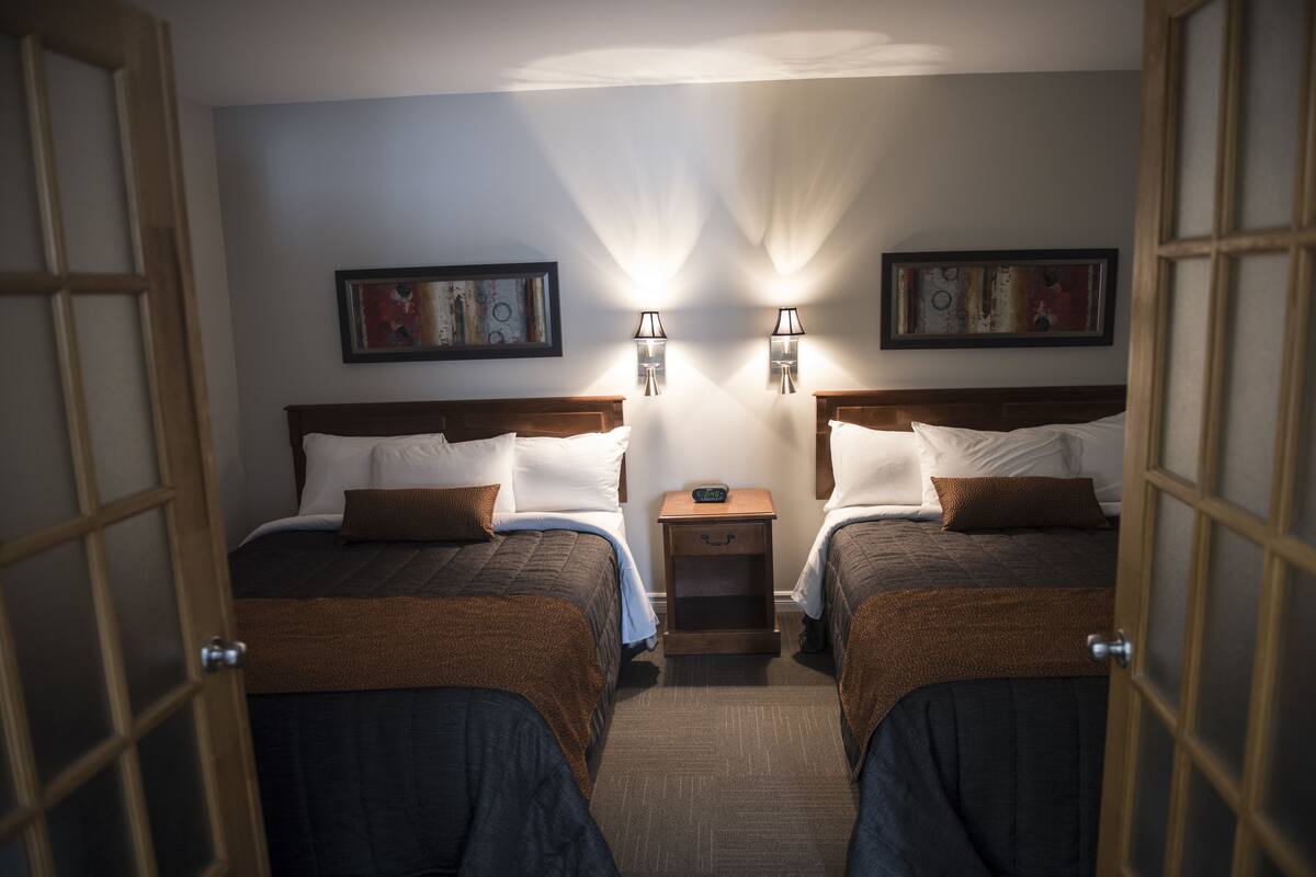 A hotel room with two beds, each with a brown blanket, white pillows, and a small bedside table with a lamp. There are two framed pictures on the wall.