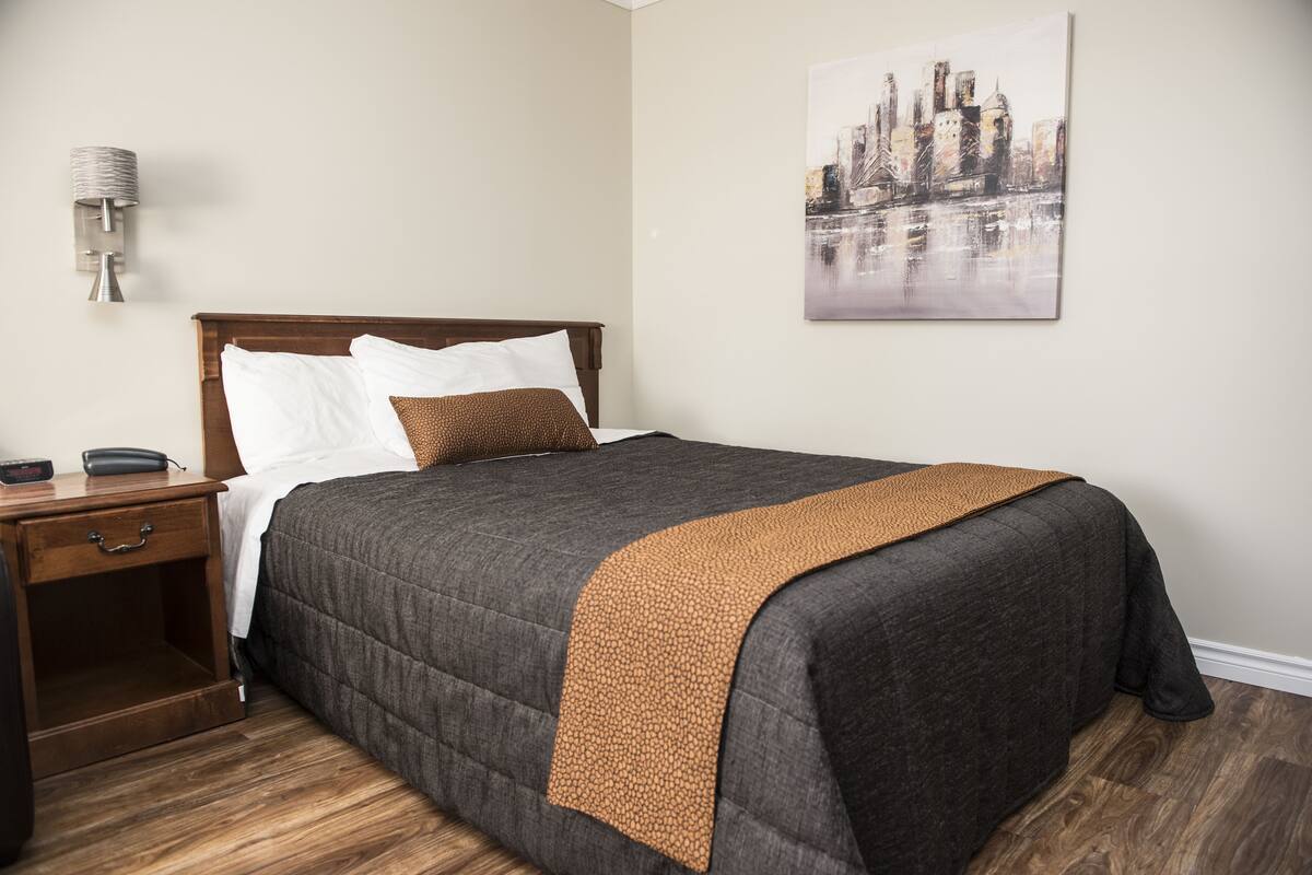 A neatly made bed with a dark grey quilt and a brown throw, a wooden nightstand with a lamp, and a cityscape painting on the wall.