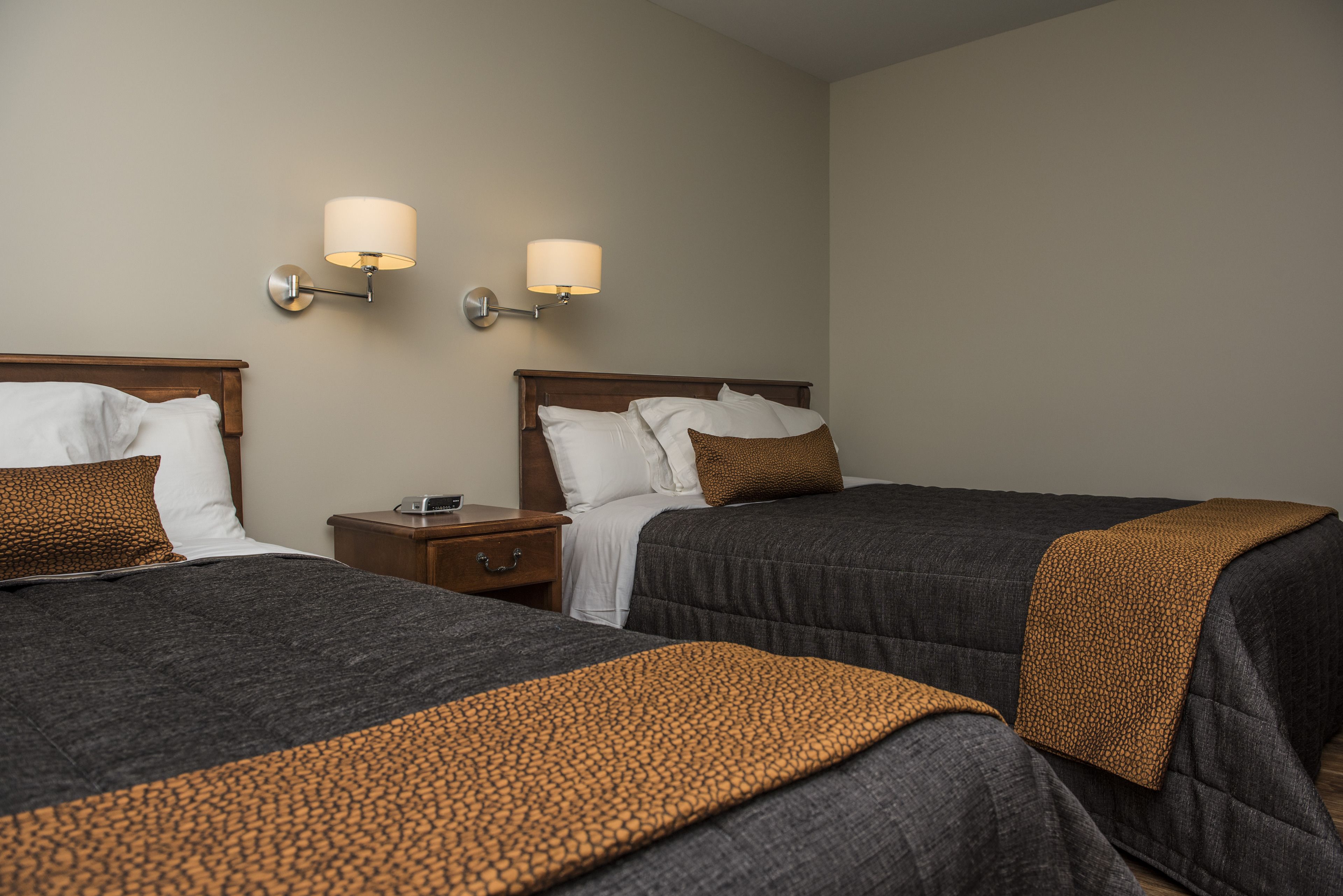 Une chambre d’hôtel avec deux lits, chacun recouvert d’une couverture grise foncé et d’un plaid à motifs. Deux lampes murales sont fixées de chaque côté des lits.