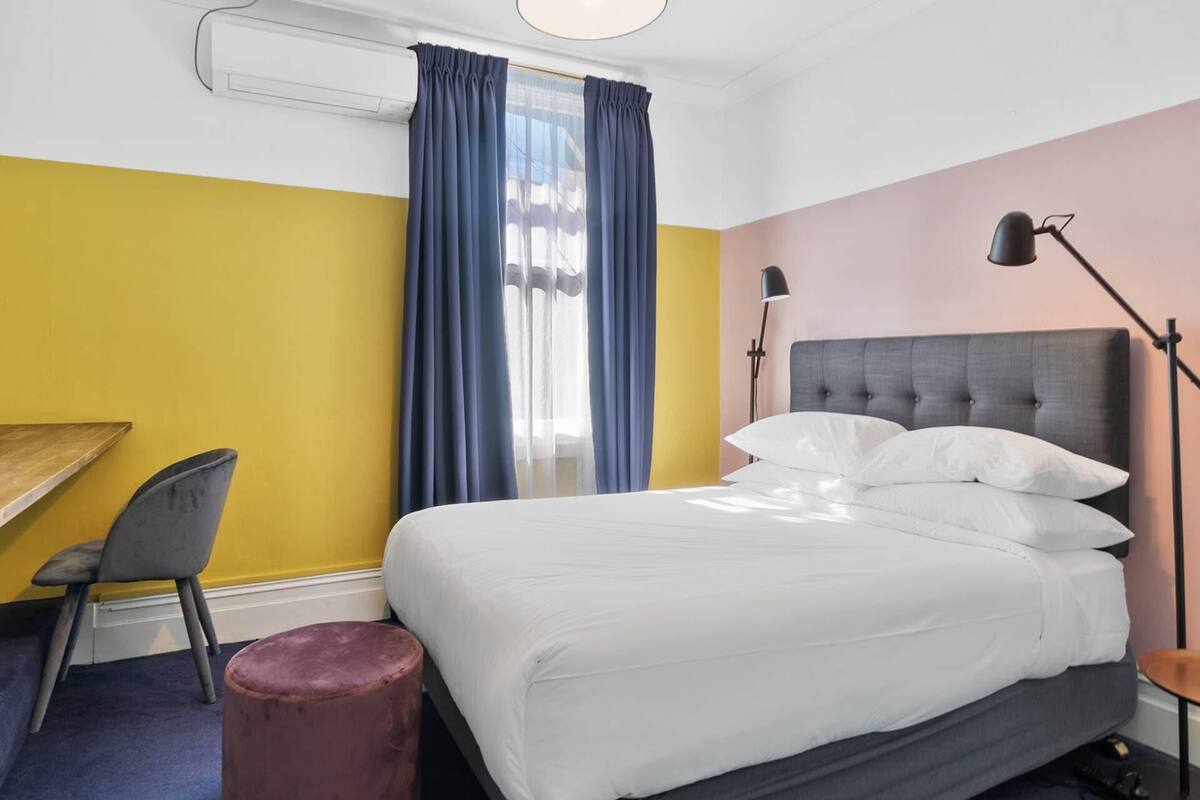 A hotel room with a bed, a desk, a chair, a lamp, and a window with curtains.