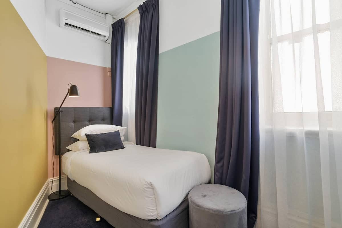 A hotel room with a bed, a bedside lamp, an air conditioning unit, and a window with curtains.