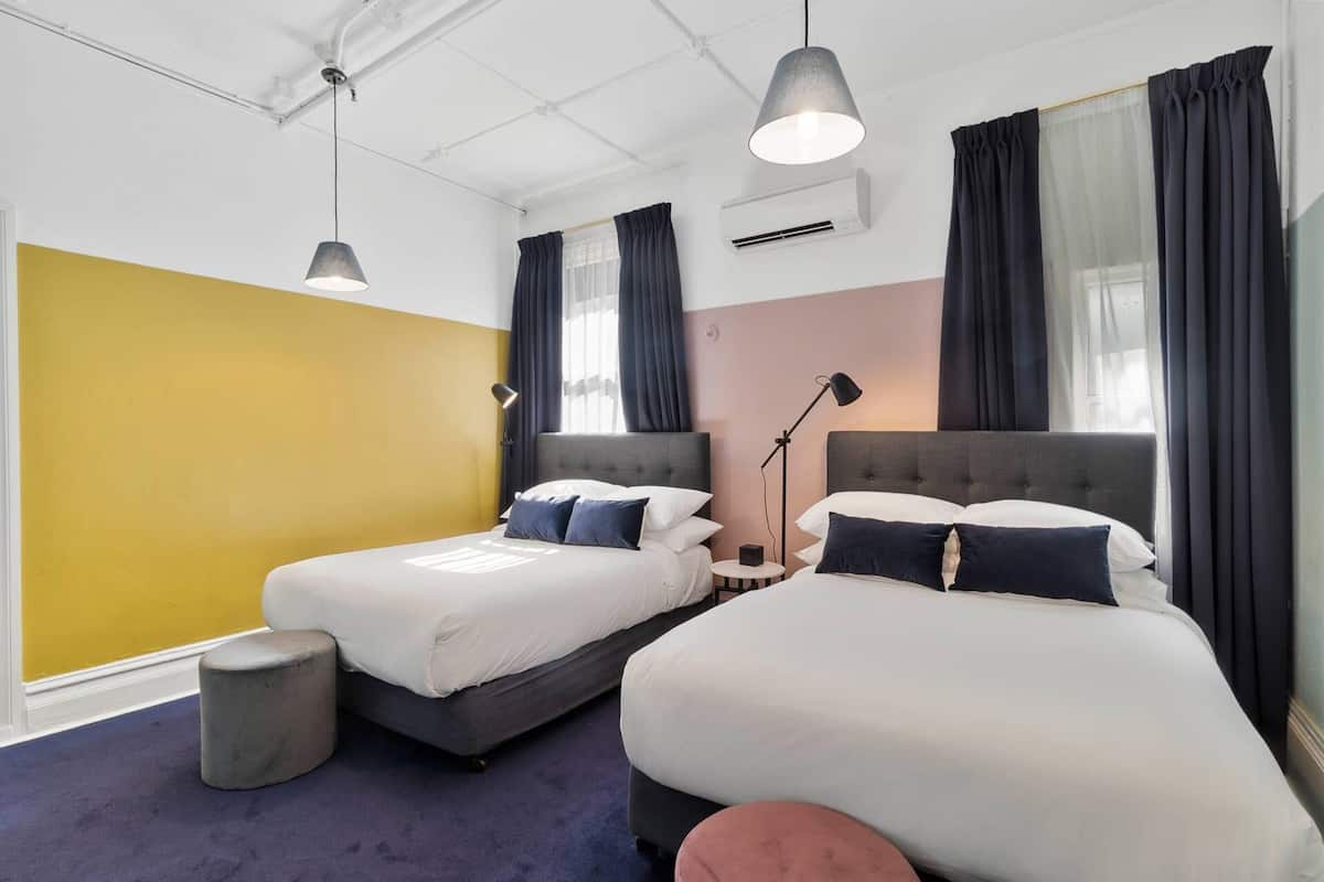 A hotel room with two beds, a small stool, a standing lamp, and an air conditioning unit.