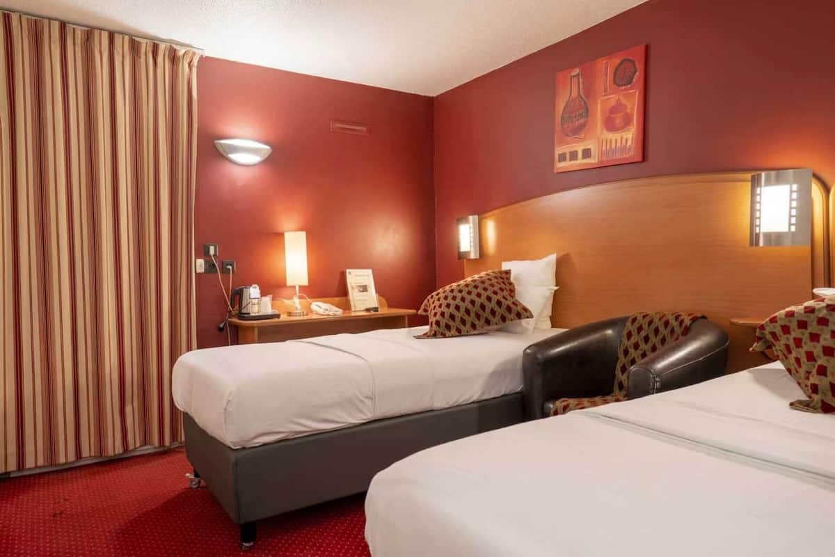 Ein Hotelzimmer mit zwei Betten, einem Holzkopfende, einer roten Wand, einem Vorhang, einem Nachttisch mit Lampe und einem Gemälde.