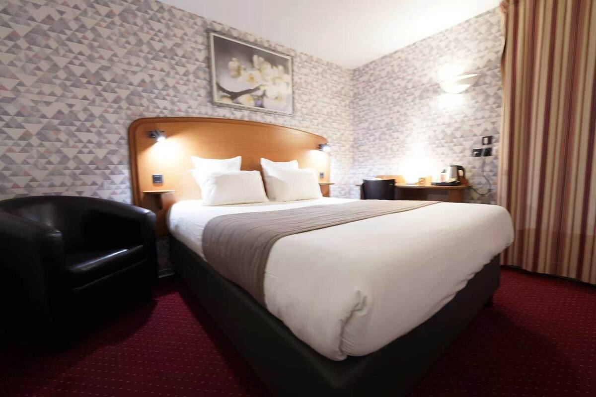 Ein Hotelzimmer mit einem Bett, einem Sessel, einem Nachttisch mit Lampe und einem Bild an der Wand.
