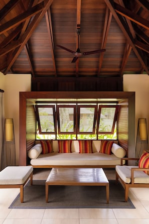 A room with a wooden ceiling, a sofa with colorful cushions, a coffee table, and a view through a window with blinds.