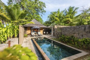 Un área de piscina con sillas de descanso, una zona de estar cubierta pequeña y abundante vegetación.
