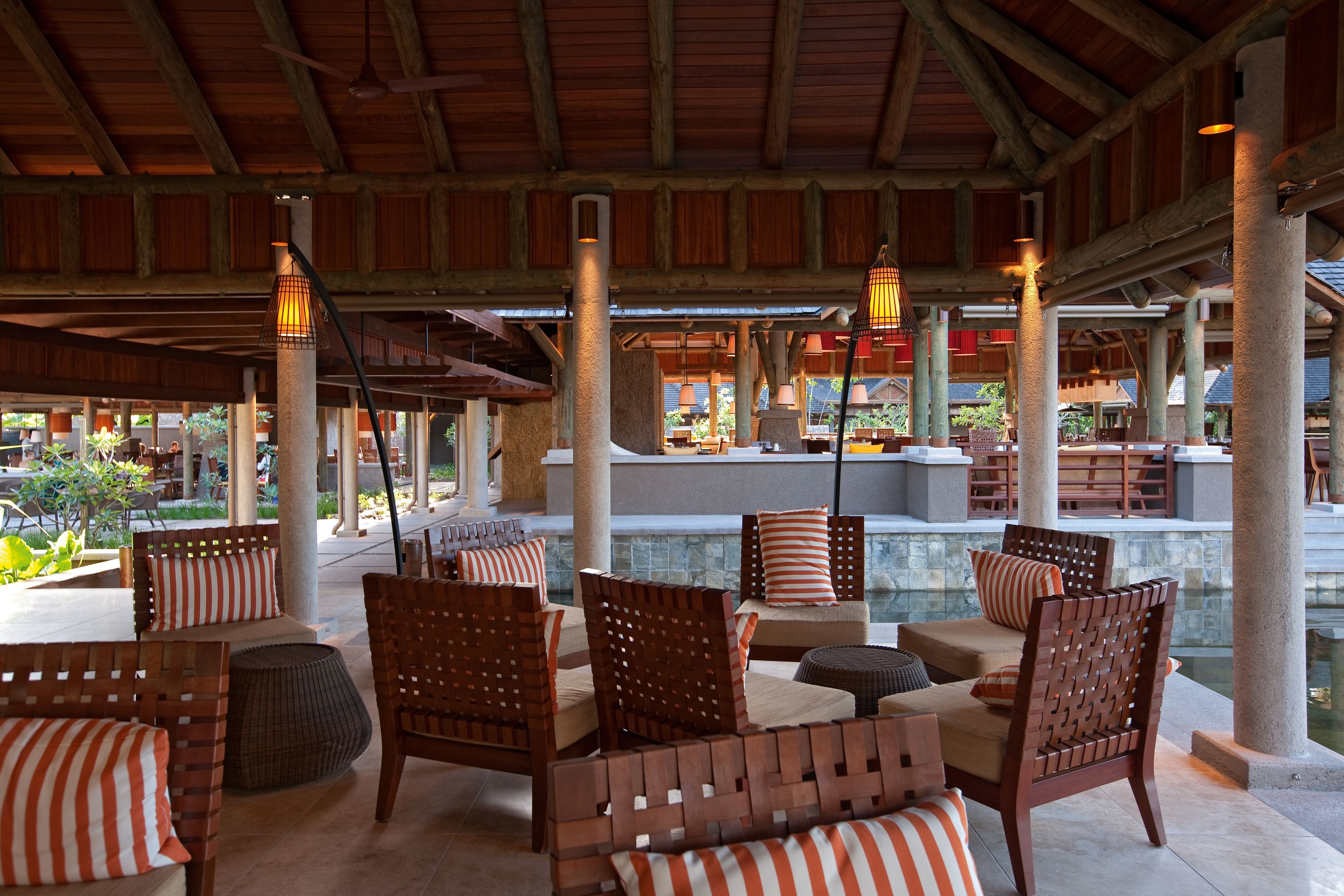 A covered outdoor seating area with wooden furniture and pillars, a pool, and a stone wall in the background.