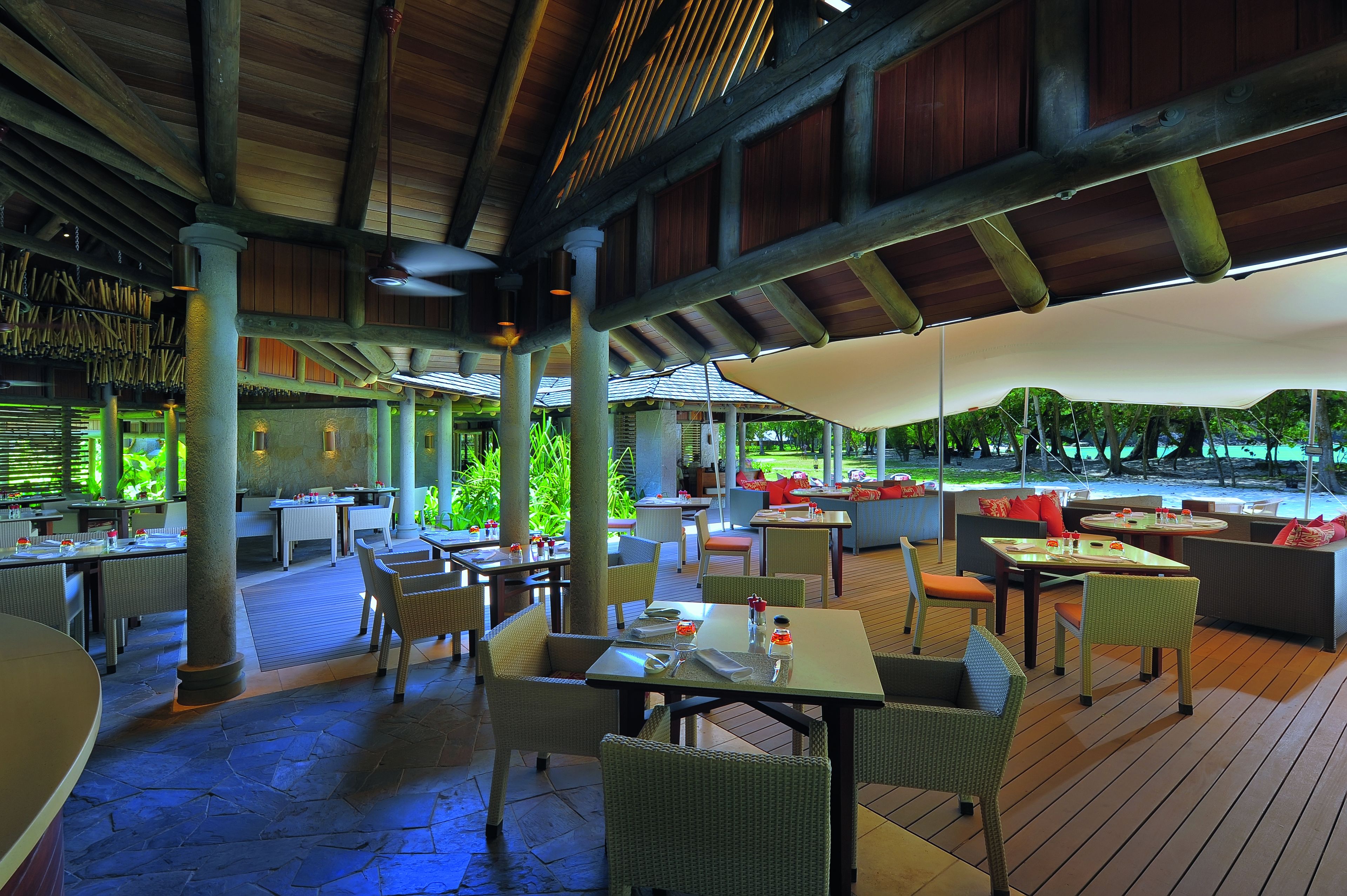 An outdoor dining area with tables and chairs, covered by large umbrellas, and surrounded by greenery.
