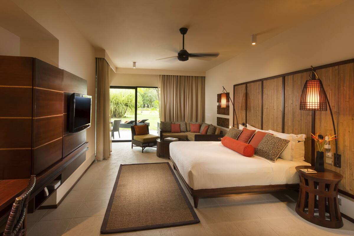 Amplia habitación de hotel con una cama grande, un televisor de pantalla plana, ventilador de techo y vistas al jardín a través de la puerta corrediza de vidrio.